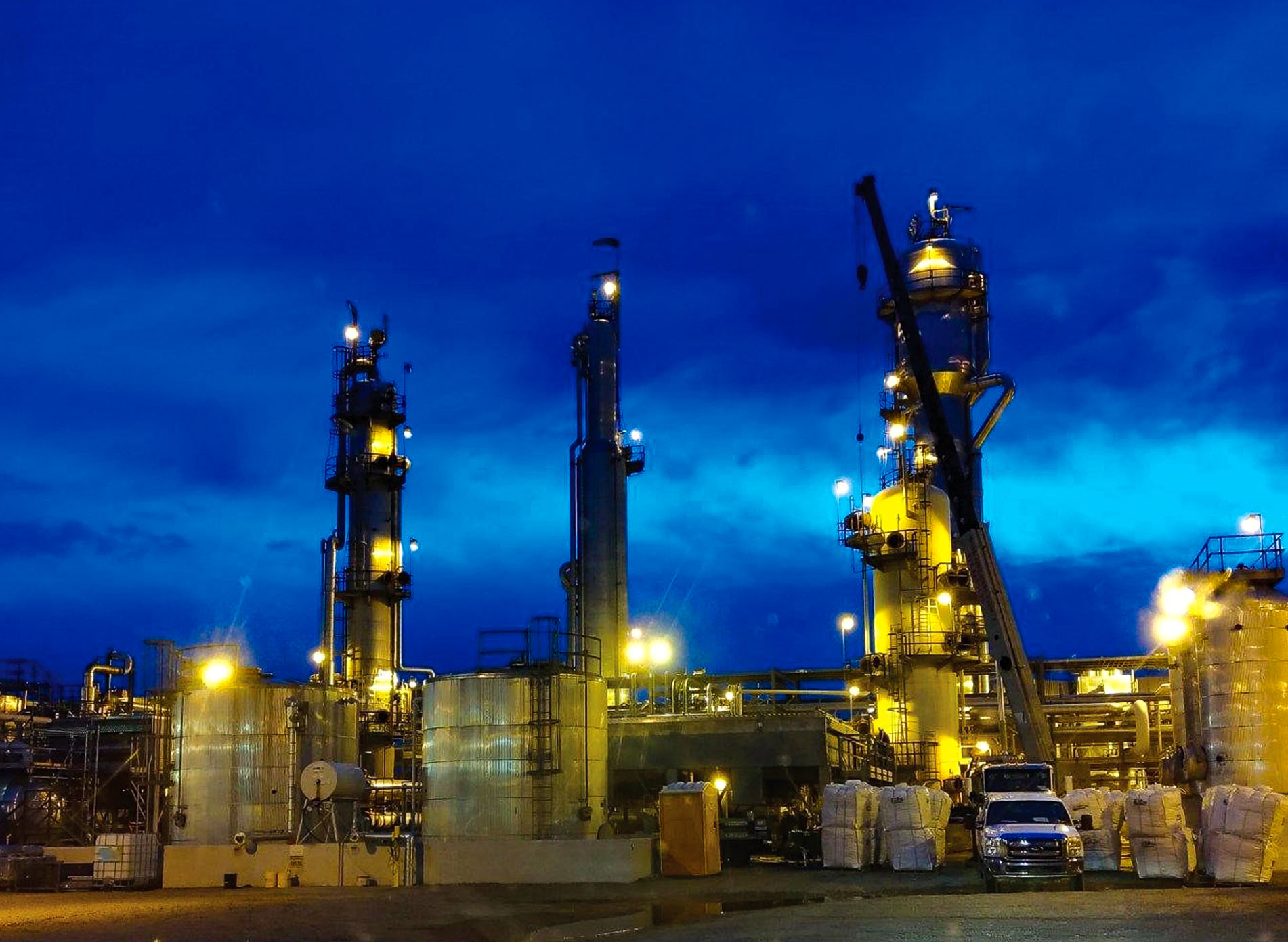 Oil refinery at night with illuminated towers and industrial equipment, showcasing active oilfield operations under a deep blue sky.
